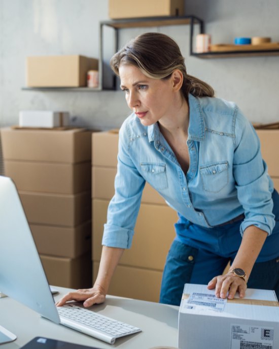 A business owner at her desk checking an order on screen while holding a shipping box